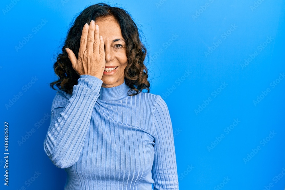 Middle age hispanic woman wearing casual clothes covering one eye with hand, confident smile on face and surprise emotion. Middle age hispanic woman wearing casual clothes covering one eye with hand, confident smile on face and surprise emotion.