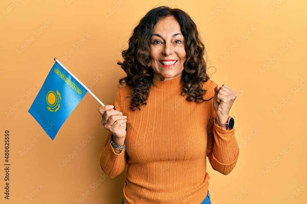 Middle age hispanic woman holding kazakhstan flag screaming proud, celebrating victory and success very excited with raised arm Middle age hispanic woman holding kazakhstan flag screaming proud, celebrating victory and success very excited with raised arm