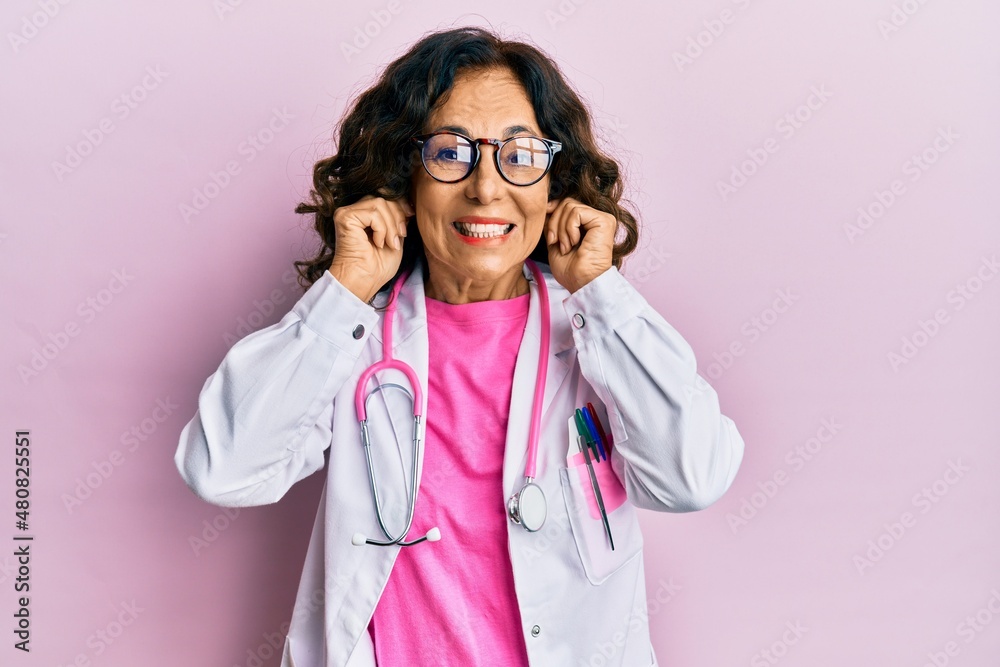 Middle age hispanic woman wearing doctor uniform and glasses covering ears with fingers with annoyed expression for the noise of loud music. deaf concept. Middle age hispanic woman wearing doctor uniform and glasses covering ears with fingers with annoyed expression for the noise of loud music. deaf concept.