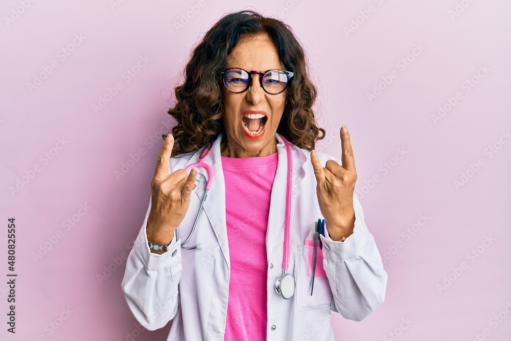 Middle age hispanic woman wearing doctor uniform and glasses shouting with crazy expression doing rock symbol with hands up. music star. heavy concept.