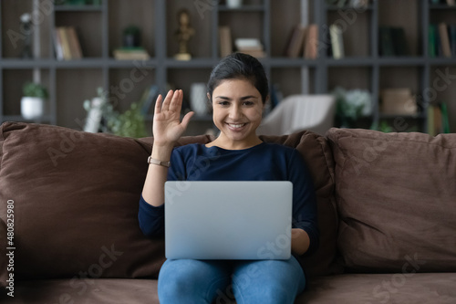 Smiling attractive young Indian ethnicity woman waving hand, starting video call meeting uon computer. Happy female blogger recording video, streaming online for social network at home. photo