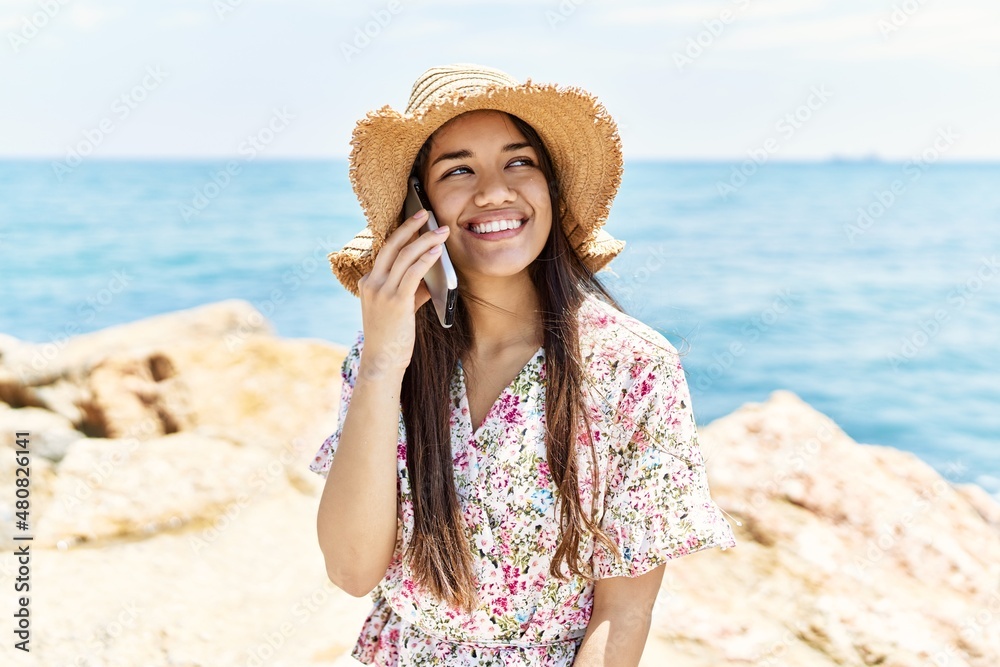 Young latin girl wearing summer hat talking on the smartphone at the beach.
