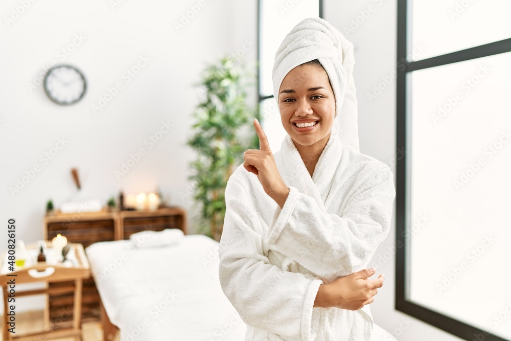 Young brunette woman wearing towel and bathrobe standing at beauty center with a big smile on face, pointing with hand and finger to the side looking at the camera.