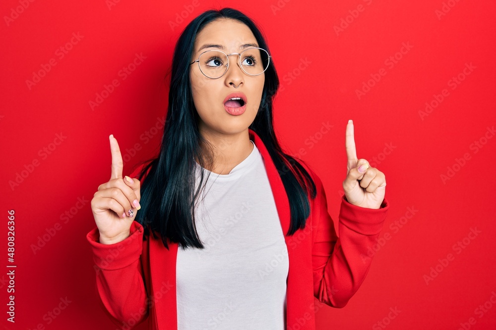 Beautiful hispanic woman with nose piercing wearing casual look and glasses amazed and surprised looking up and pointing with fingers and raised arms. Beautiful hispanic woman with nose piercing wearing casual look and glasses amazed and surprised looking up and pointing with fingers and raised arms.