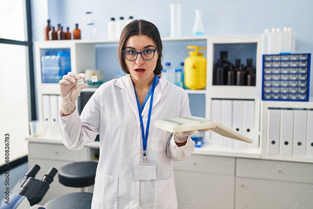 Young hispanic woman working at scientist laboratory with blood samples in shock face, looking skeptical and sarcastic, surprised with open mouth