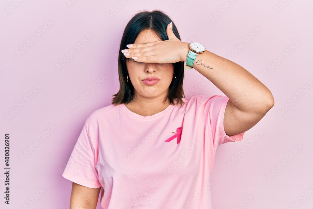 Young hispanic woman wearing pink cancer ribbon on t shirt covering eyes with hand, looking serious and sad. sightless, hiding and rejection concept