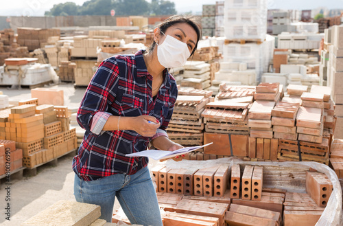 Female worker in protective face mask checking quantity of red bricks in warehouse of building materials