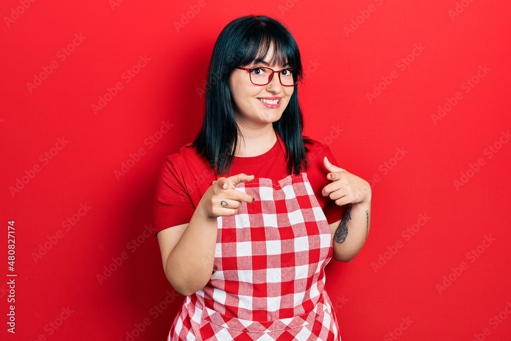 Young hispanic woman wearing cook apron and glasses pointing fingers to camera with happy and funny face. good energy and vibes. Young hispanic woman wearing cook apron and glasses pointing fingers to camera with happy and funny face. good energy and vibes.