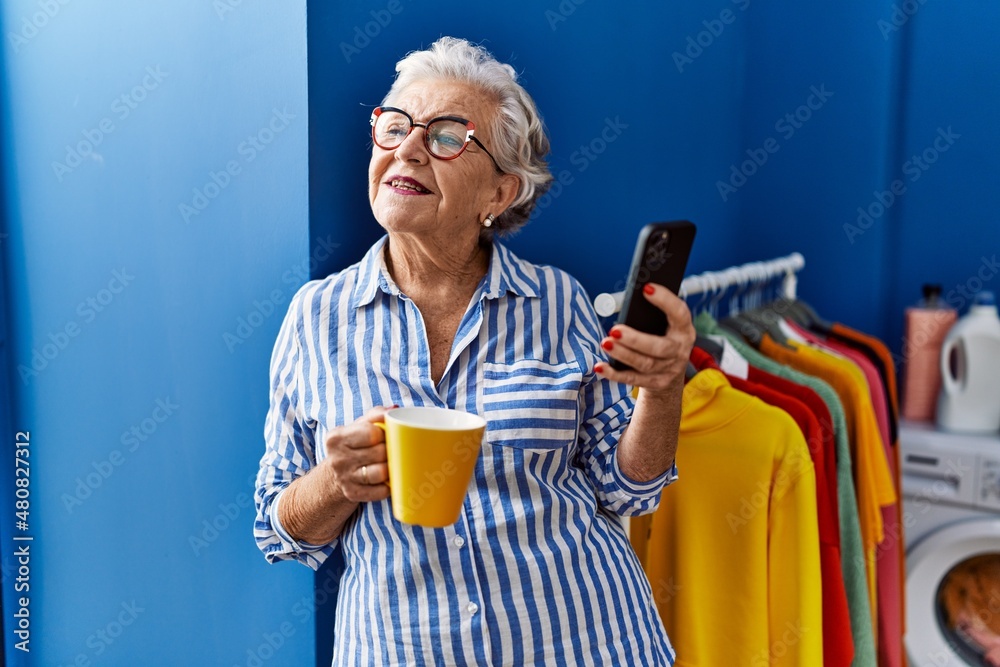 Senior grey-haired woman using smartphone drinking coffee at laundry room Senior grey-haired woman using smartphone drinking coffee at laundry room