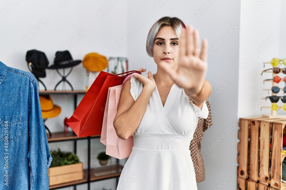 Young beautiful woman holding shopping bags at retail shop with open hand doing stop sign with serious and confident expression, defense gesture Young beautiful woman holding shopping bags at retail shop with open hand doing stop sign with serious and confident expression, defense gesture