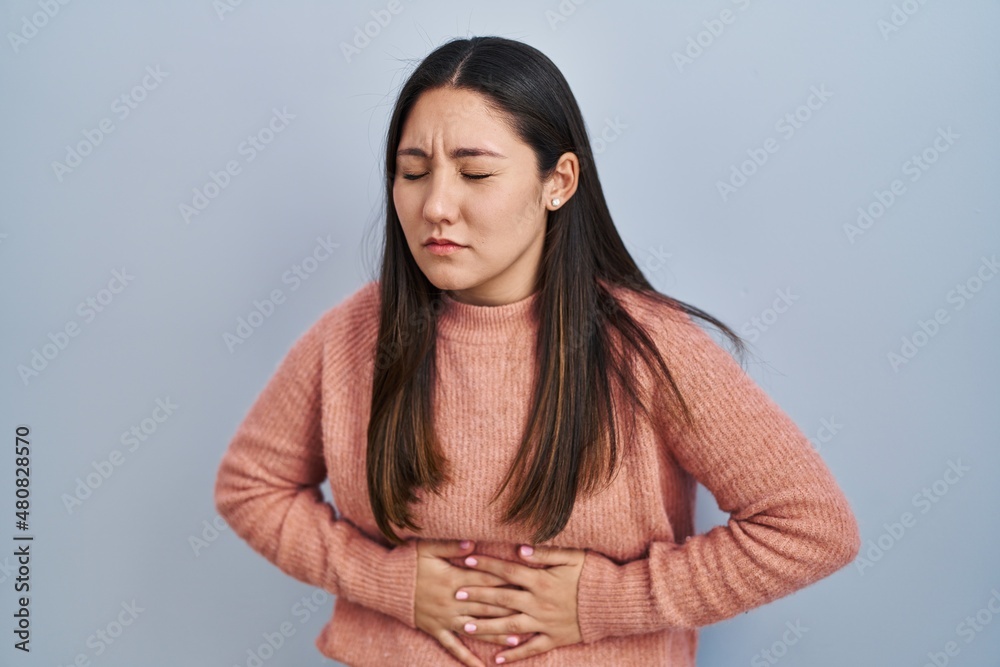Young latin woman standing over blue background with hand on stomach because indigestion, painful illness feeling unwell. ache concept. Young latin woman standing over blue background with hand on stomach because indigestion, painful illness feeling unwell. ache concept.