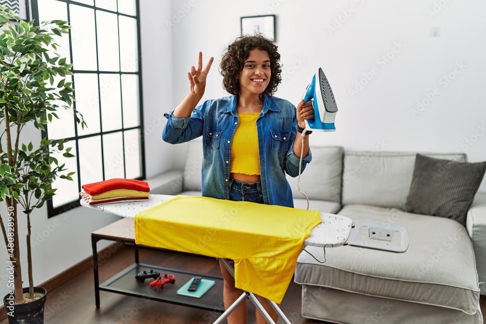 Young hispanic woman ironing clothes at home smiling looking to the camera showing fingers doing victory sign. number two.