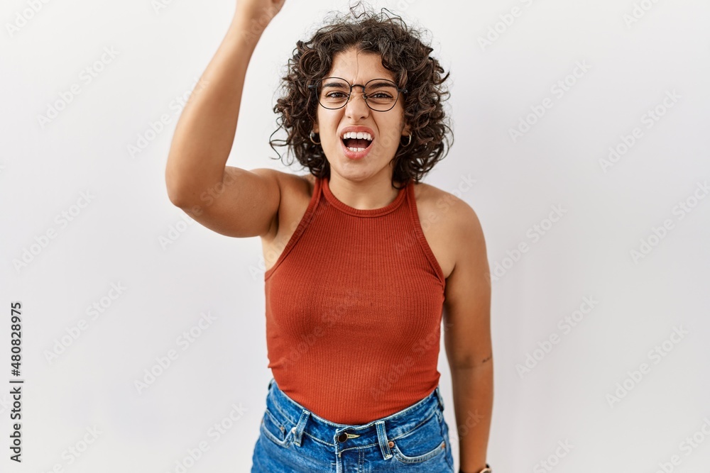Young hispanic woman wearing glasses standing over isolated background angry and mad raising fist frustrated and furious while shouting with anger. rage and aggressive concept. Young hispanic woman wearing glasses standing over isolated background angry and mad raising fist frustrated and furious while shouting with anger. rage and aggressive concept.