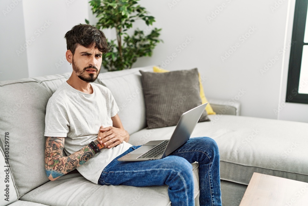 Hispanic man with beard sitting on the sofa with hand on stomach because indigestion, painful illness feeling unwell. ache concept.