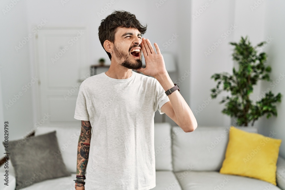Hispanic man with beard at the living room at home shouting and screaming loud to side with hand on mouth. communication concept. Hispanic man with beard at the living room at home shouting and screaming loud to side with hand on mouth. communication concept.