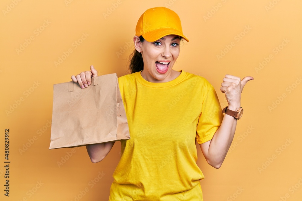 Young hispanic woman holding take away paper bag pointing thumb up to the side smiling happy with open mouth