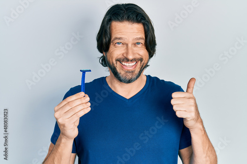 Middle age caucasian man holding razor smiling happy and positive, thumb up doing excellent and approval sign