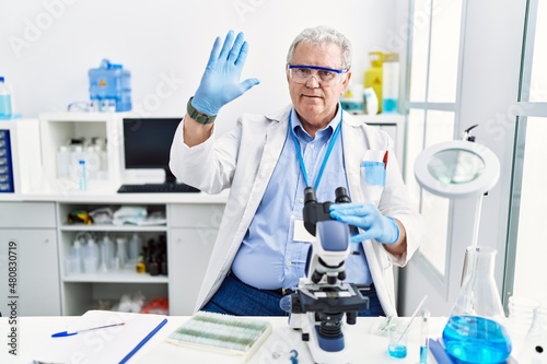 Senior caucasian man working at scientist laboratory waiving saying hello happy and smiling  friendly welcome gesture