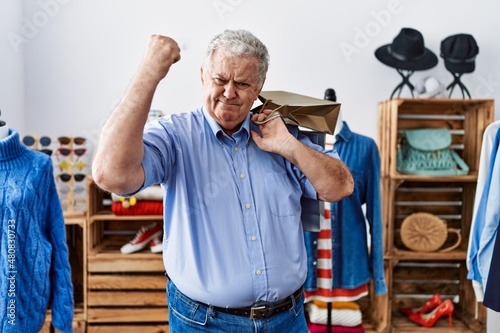 Senior man with grey hair holding shopping bags at retail shop annoyed and frustrated shouting with anger, yelling crazy with anger and hand raised