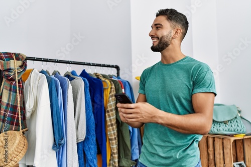 Young arab man customer using smartphone standing by clothes rack at clothing store