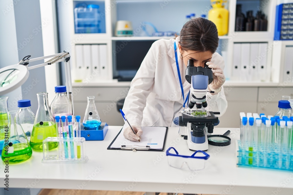 Fototapeta premium Young hispanic woman wearing scientist uniform using microscope and writing on clipboard at laboratory