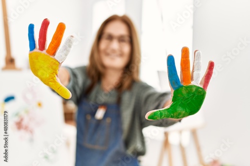 Middle age artist woman smiling happy showing painted hands colorful paint at art studio.