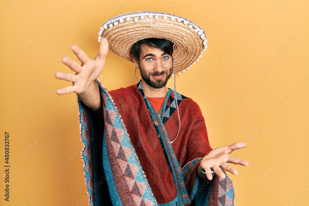 Young hispanic man holding mexican hat looking at the camera smiling with open arms for hug. cheerful expression embracing happiness.