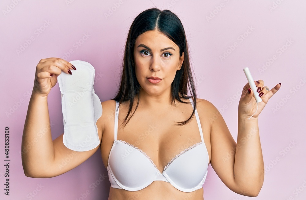 Beautiful brunette woman holding sanitary pad and cotton tampon relaxed with serious expression on face. simple and natural looking at the camera. Beautiful brunette woman holding sanitary pad and cotton tampon relaxed with serious expression on face. simple and natural looking at the camera.
