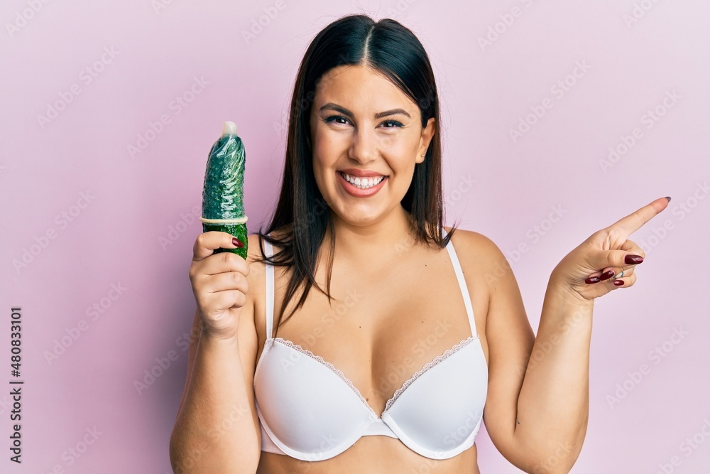 Beautiful brunette woman holding condom on cucumber for sex education smiling happy pointing with hand and finger to the side