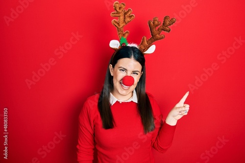 Young hispanic woman wearing deer christmas hat and red nose with a big smile on face, pointing with hand finger to the side looking at the camera.