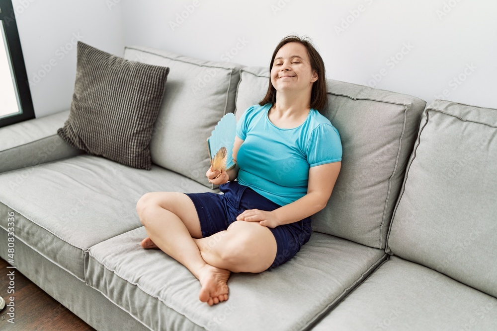 Brunette woman with down syndrome sitting on the sofa using hand fan at the living room Brunette woman with down syndrome sitting on the sofa using hand fan at the living room