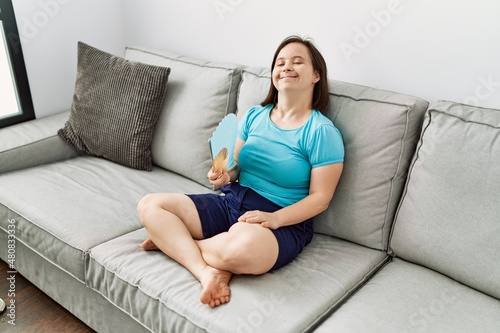 Brunette woman with down syndrome sitting on the sofa using hand fan at the living room