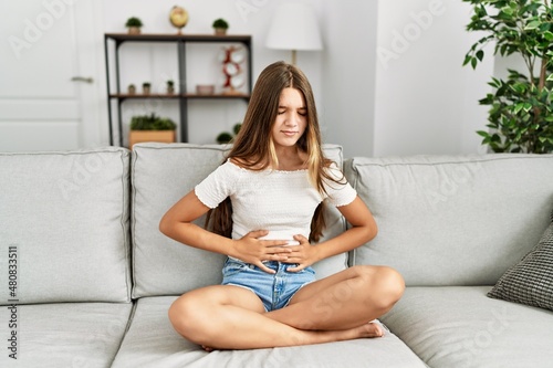 Young brunette teenager sitting on the sofa at home with hand on stomach because indigestion, painful illness feeling unwell. ache concept.