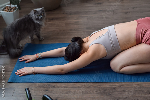 Young girl on yoga mat in childs pose working out at home with cat walking by