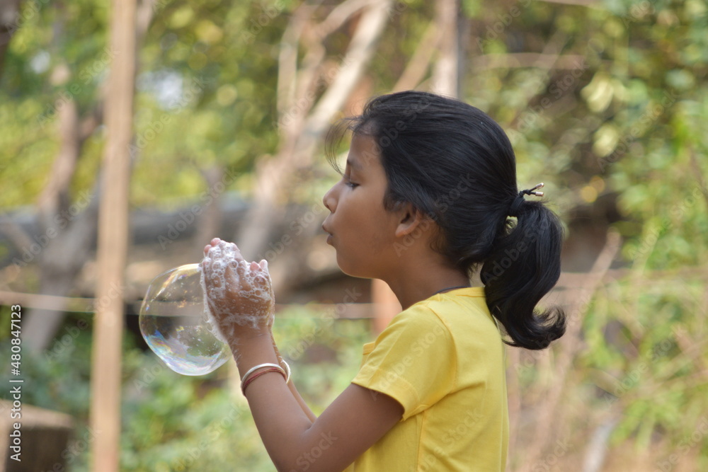 A girl kid making soap bubbles wearing yellow tshirt in backyard garden selectively focused with blurr background A girl kid making soap bubbles wearing yellow tshirt in backyard garden selectively focused with blurr background
