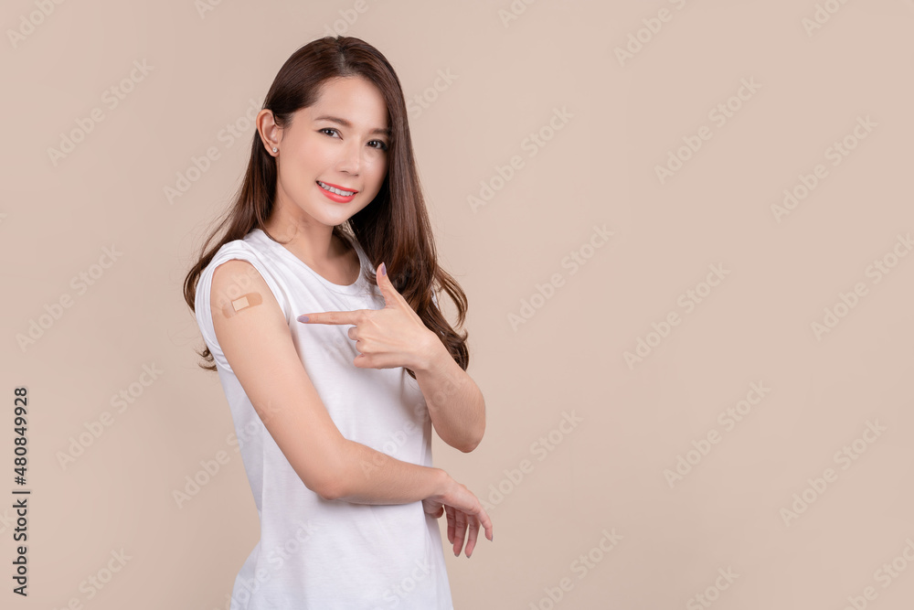 Obraz premium Coronavirus vaccination protects lives. Young beautiful asian woman pointing at adhesive bandage on her arm, She getting vaccinated against covid-19 on beige background.