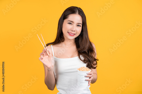 Beautiful Asian woman holding a cup of spicy instant noodles on a yellow background. Fast food concept.