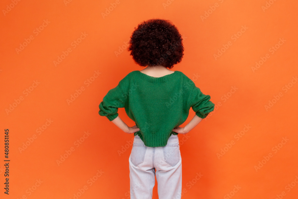 Back view portrait of woman with Afro hairstyle wearing green casual style sweater standing posing backwards, keeping hands on hips. Indoor studio shot isolated on orange background. Back view portrait of woman with Afro hairstyle wearing green casual style sweater standing posing backwards, keeping hands on hips. Indoor studio shot isolated on orange background.