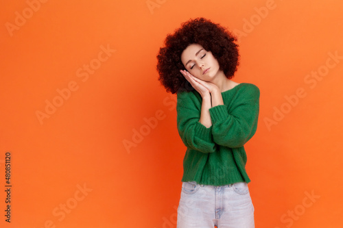 Portrait of sleepy bored woman with Afro hairstyle wearing green casual style sweater leaning head on hands, keeps eyes closed, copy space. Indoor studio shot isolated on orange background. © khosrork