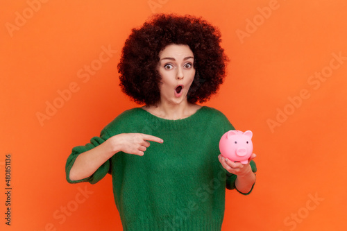 Portrait of shocked woman with Afro hairstyle wearing green casual style sweater standing pointing at piggy bank in her hand, advantageous bank offer. Indoor studio shot isolated on orange background. © khosrork