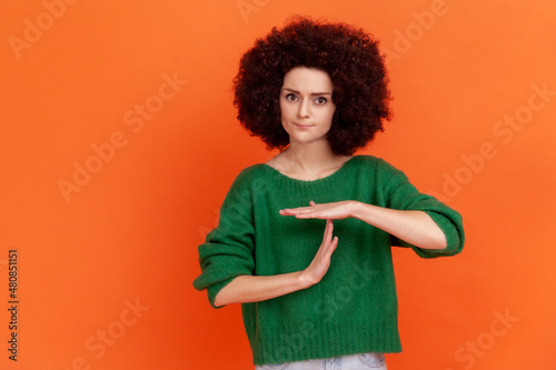 Young adult woman with Afro hairstyle wearing green casual style sweater showing time out hand gesture, looking at camera, worried about deadline. Indoor studio shot isolated on orange background. photo