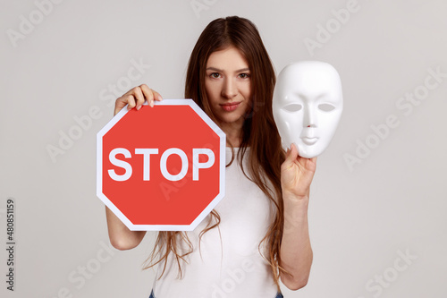 Portrait of serious dark haired woman holding white mask with unknown face and red traffic sign, looking at camera, wearing white T-shirt. Indoor studio shot isolated on gray background. photo