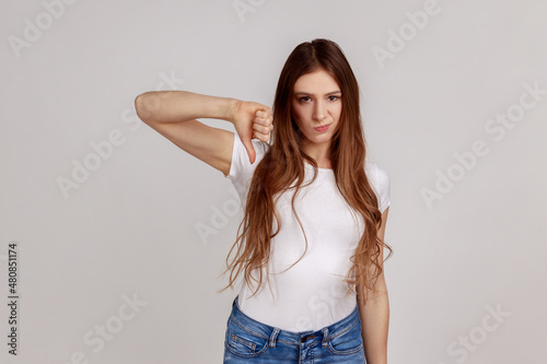 Portrait of dark haired unhappy unsatisfied woman showing thumb down, dislike gesture, negative emotions, bad rating, wearing white T-shirt. Indoor studio shot isolated on gray background.