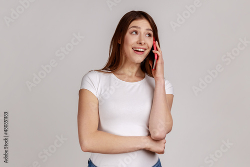 Beautiful young adult woman talking phone and smiling, pleasant conversation with boyfriend, making appointment or date, wearing white T-shirt. Indoor studio shot isolated on gray background.