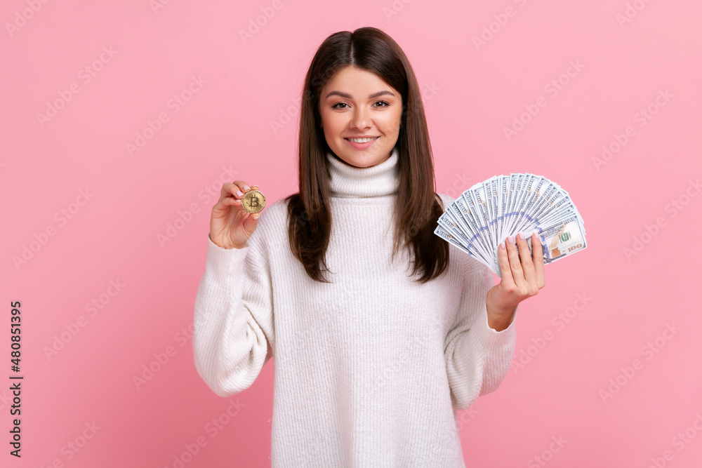 Optimistic dark haired woman holding bitcoin and big fan of dollar bills, exchange crypto currency, wearing white casual style sweater. Indoor studio shot isolated on pink background. Optimistic dark haired woman holding bitcoin and big fan of dollar bills, exchange crypto currency, wearing white casual style sweater. Indoor studio shot isolated on pink background.