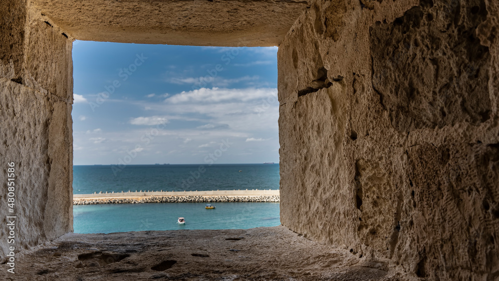 Through a square opening in the thick wall of the ancient Citadel of Qaitbay in Alexandria, you can see the blue sky, the turquoise Sea, boats on the water.  The rough texture of the stone is visible.