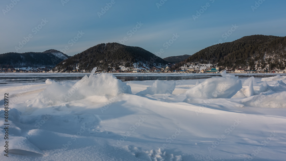 Bizarre figures of icy snow formed on the surface of a frozen lake. In the distance, on the shore, the village is visible. Wooded mountains against a blue sky. Baikal