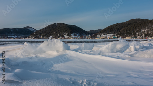 Bizarre figures of icy snow formed on the surface of a frozen lake. In the distance, on the shore, the village is visible. Wooded mountains against a blue sky. Baikal © Вера 