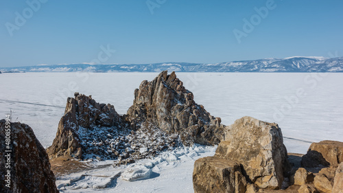 A picturesque granite double-headed rock on the background of a frozen lake. Cracks on slopes devoid of vegetation. A mountain range in the distance. Boulders in the foreground. Blue sky. Baikal