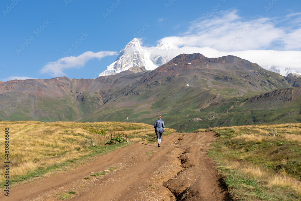 A woman walking on a panoramic hiking trail leading to the Koruldi Lakes in the Greater Caucasus Mountain Range in Georgia, Upper Svanet Region. Panoramic view on the Ushba mountain peaks. Wanderlust.